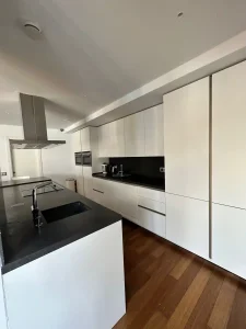 Cocina moderna blanca con isla central, encimera oscura, campana extractora y suelo de madera.