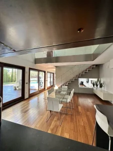 Salón comedor de concepto abierto con cocina, escalera de diseño, grandes ventanales y vista a la piscina.