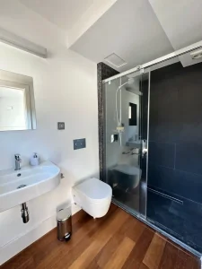 Baño moderno con lavabo blanco, inodoro suspendido, ducha negra acristalada y ventana alta.