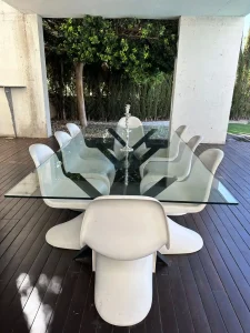 Mesa exterior de cristal con sillas blancas sobre tarima de madera junto a la piscina.