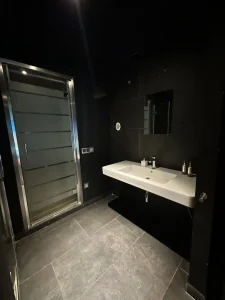 Baño moderno en tonos negros con lavabo doble, ducha acristalada y acabados oscuros.