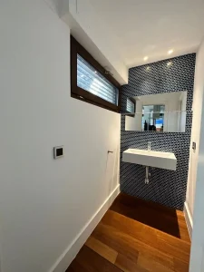 Baño con mosaico oscuro, lavabo suspendido, espejo grande y ventana alta con luz natural.