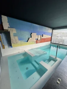 Piscina interior con zona de jacuzzi, agua turquesa y mural decorativo en la pared principal.