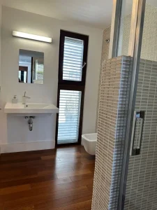 Baño luminoso con lavabo suspendido, ducha de mosaico, ventana vertical y entrada de luz natural.