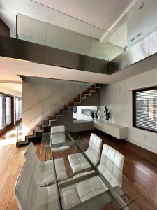 Salón comedor amplio de doble altura con mesa de cristal, sillas blancas y escalera de diseño.