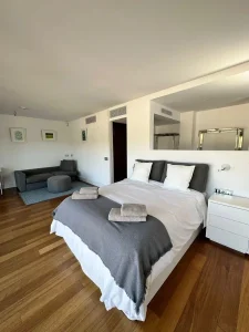 Suite amplia con cama doble, sofá gris, suelo de madera y detalles textiles preparados para huéspedes.