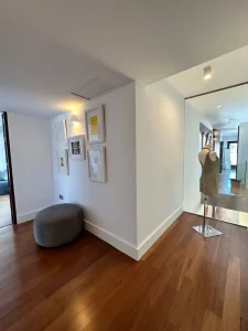 Recibidor moderno con suelo de madera, puf gris, espejo y cuadros decorativos en pared blanca.