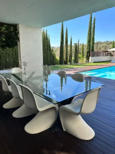 Comedor exterior con mesa de cristal y sillas blancas junto a la piscina y el jardín de cipreses.