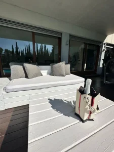 Cama exterior tipo daybed con cojines blancos sobre la terraza junto a la piscina.