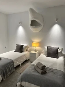 Dormitorio doble con dos camas individuales, mesita central, lámpara cálida y pared iluminada.