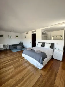 Dormitorio principal amplio con cama doble, sofá, suelo de madera y zona de estar integrada.
